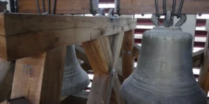 Derrière les cloches provenant de l’ancienne chapelle et le beffroi (charpente les retenant), les abat-sons